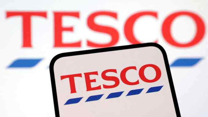 Tesco mobile logo on a smartphone, being held in front of a Tesco banner.